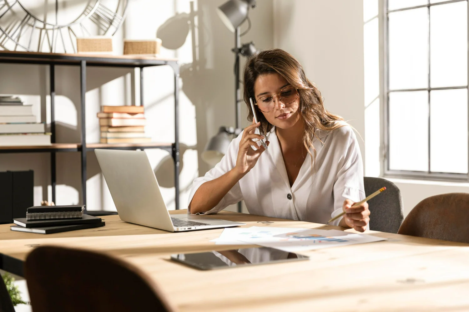 Mulher branca em uma mesa de frente para um computador e falando ao telefone enquanto tem seu serviço facilitado por um conciliador financeiro